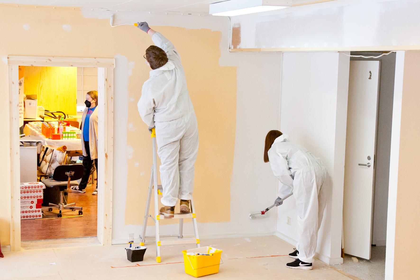 Kaksi maalaria maalaa seinää valkoiseksi suojahaalareissaan.